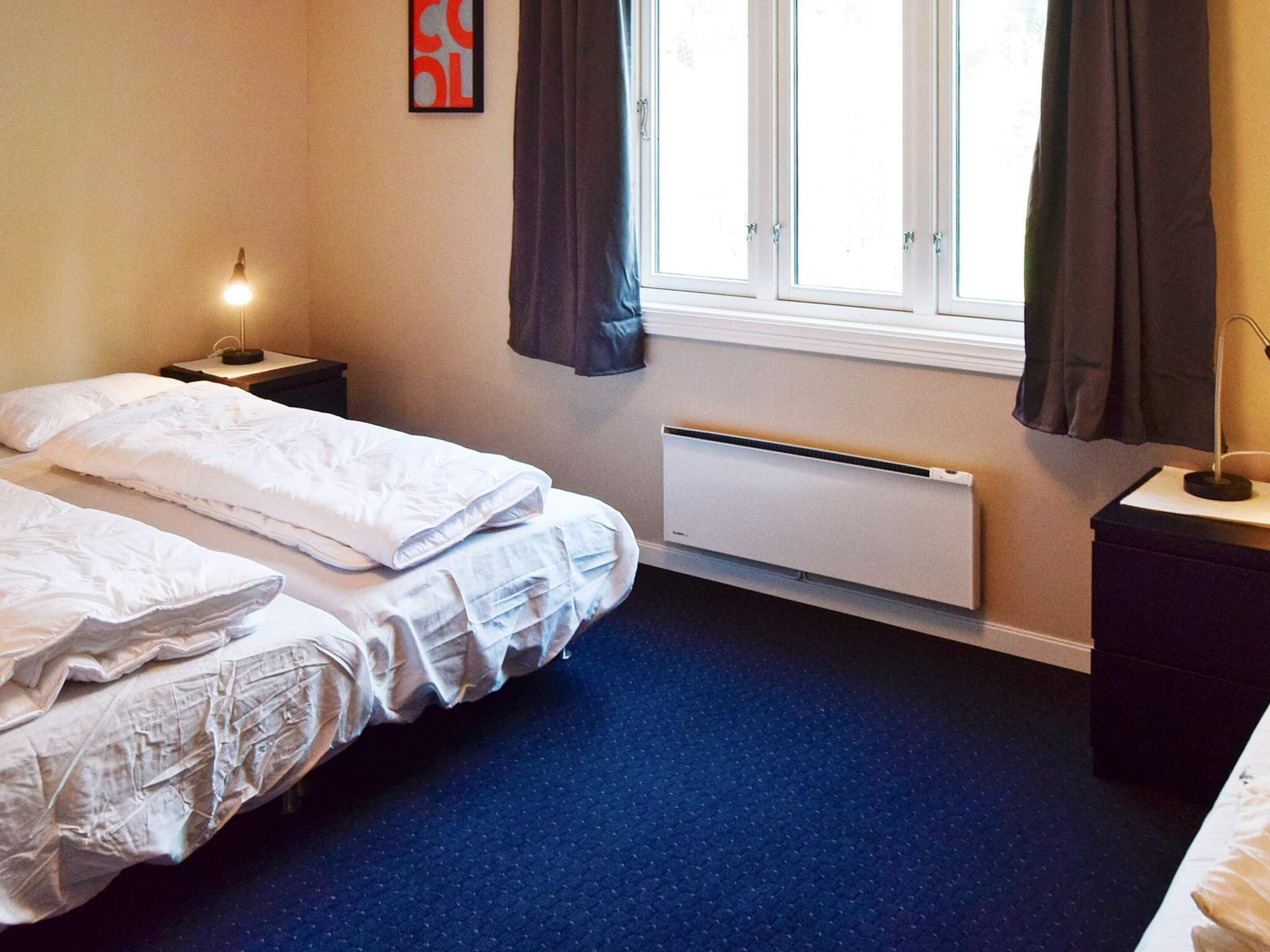 A hotel room with two beds, a window with curtains, a bedside table with a lamp, and a radiator.