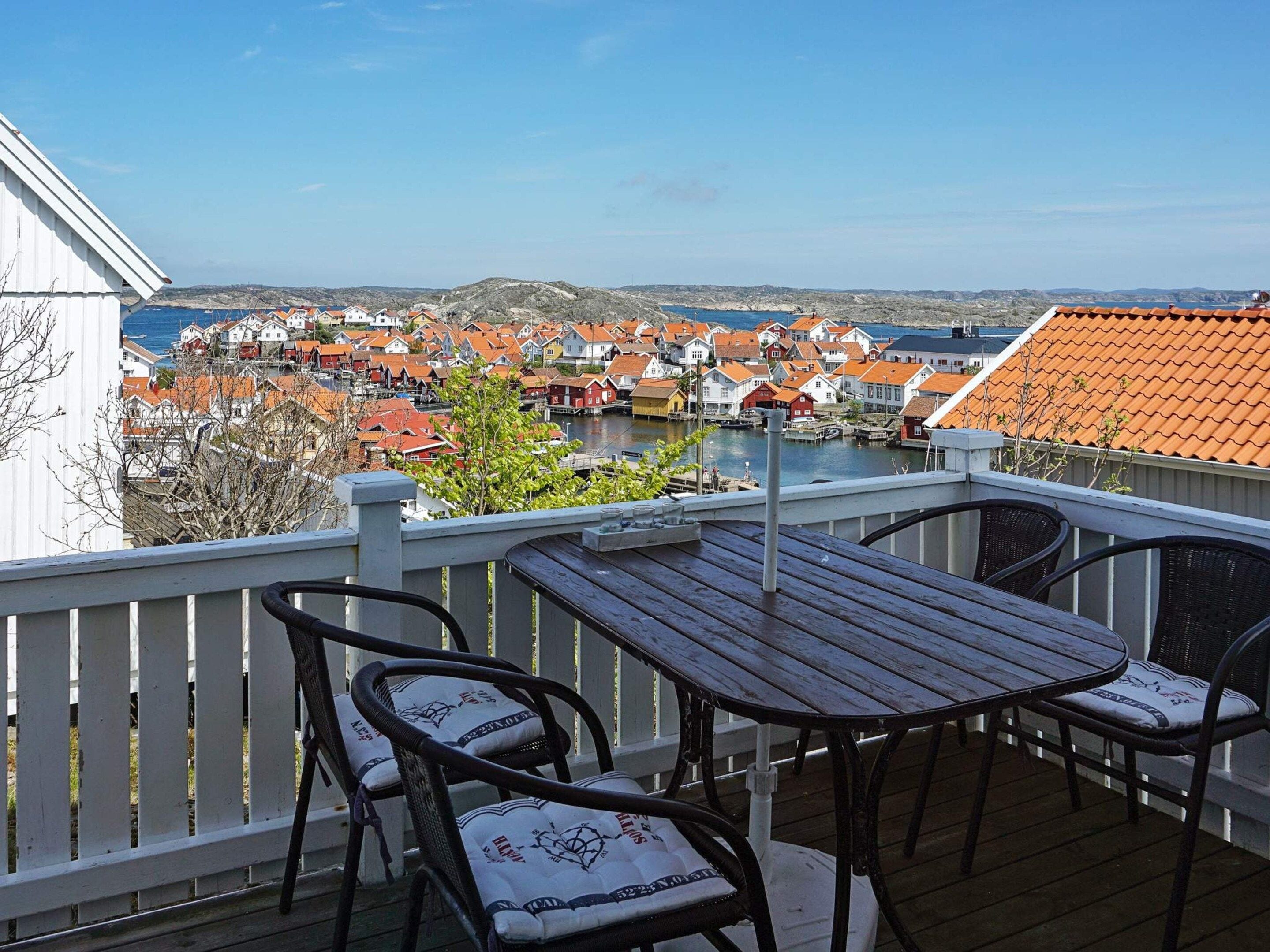 Une terrasse en bois avec une table et des chaises, offrant une vue sur une ville côtière avec des bâtiments aux toits rouges et un plan d’eau.