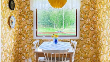 Ett rum med blommig tapet, ett träbord med stolar, ett fönster med utsikt över grönska och en gul pappersdekoration.