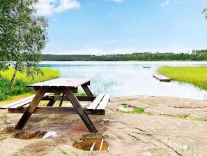 A wooden picnic table on a rocky shore with a calm lake and dock in the background. - 4 Person Holiday Home in Vimmerby (Vimmerby)