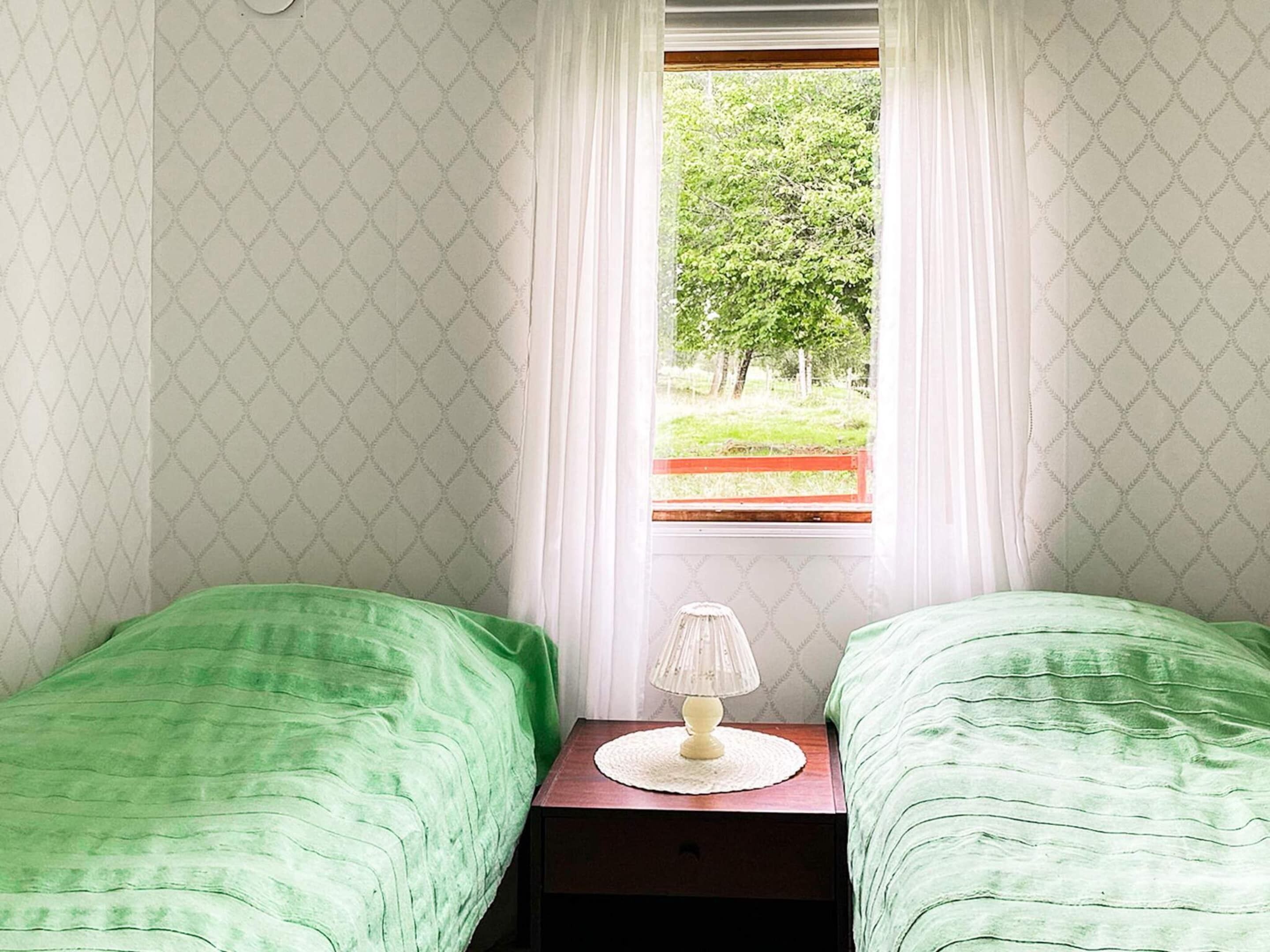 A bedroom with two single beds, a nightstand with a lamp, and a window with a view of greenery outside.