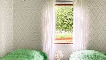 A bedroom with two single beds, a nightstand with a lamp, and a window with a view of greenery outside.