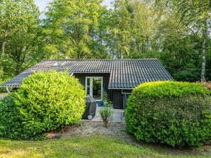 A house with a tiled roof, a door, and a window. There is a garden with bushes and a pathway leading to the house.