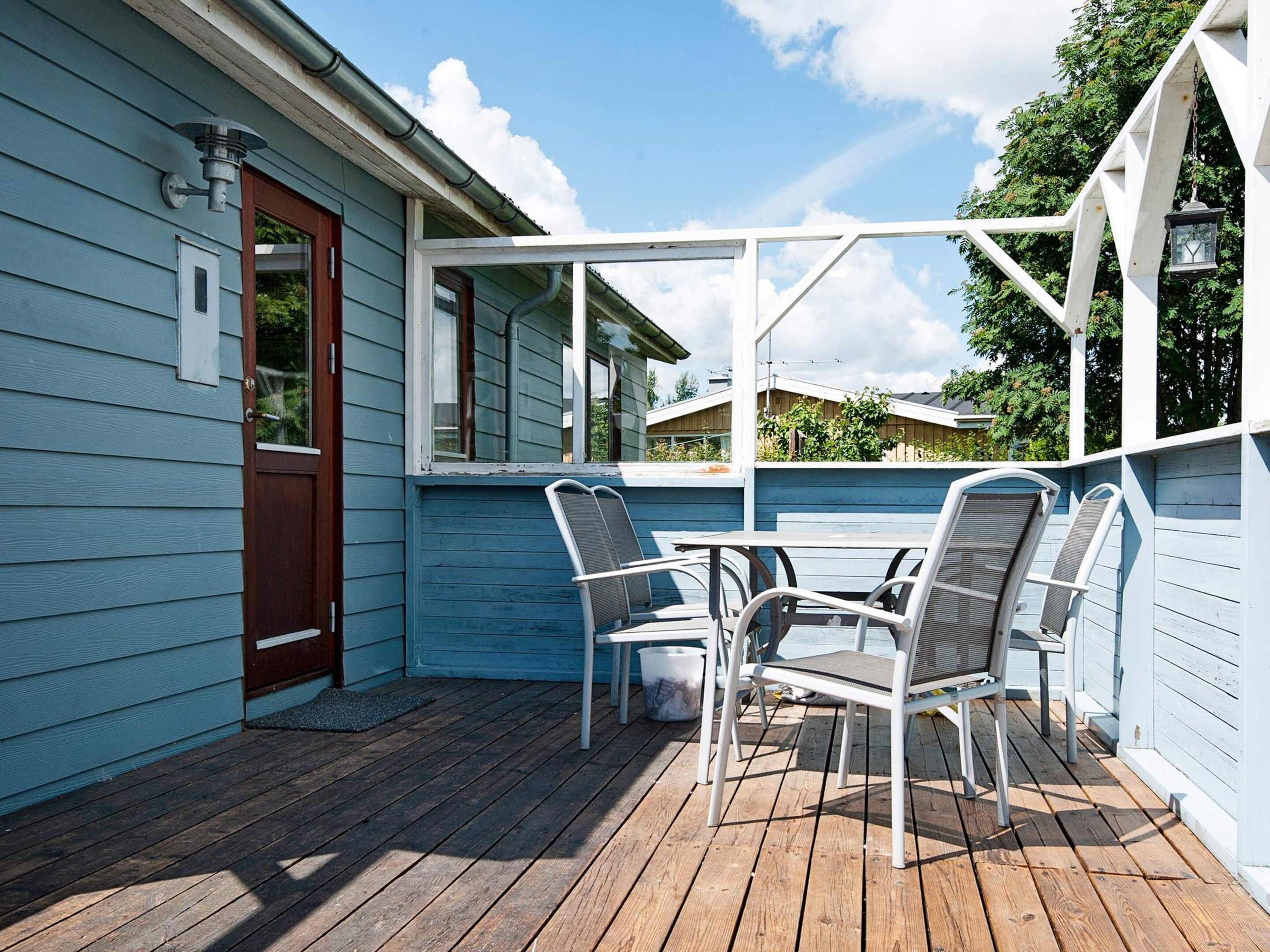 A covered outdoor seating area with a table and chairs, a wooden deck, and a house with blue siding and a brown door.