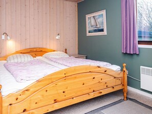 Quarto com cama de madeira, duas luminárias de apoio, um quadro com um veleiro e uma janela com cortinas roxas.