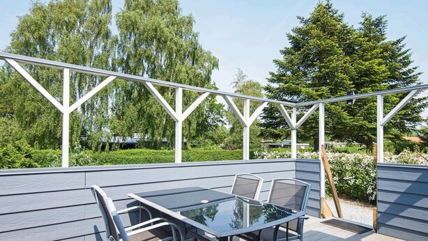 A covered outdoor deck with a glass-top table and metal chairs, surrounded by a white pergola structure. - 4 Person Holiday Home in Juelsminde (Horsens)