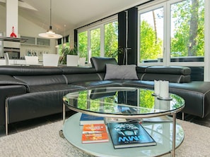 Un salon moderne avec un canapé d’angle en cuir, une table basse en verre et une vue sur l’extérieur grâce à de grandes fenêtres.
