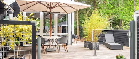 Eine Holzterrasse mit Tisch und Stühlen unter einem großen Sonnenschirm, umgeben von Blumentöpfen und einem Metallzaun.