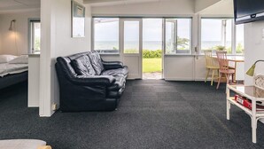 A room with a black leather sofa, a bed, a dining table with chairs, and large windows offering a view of the sea.