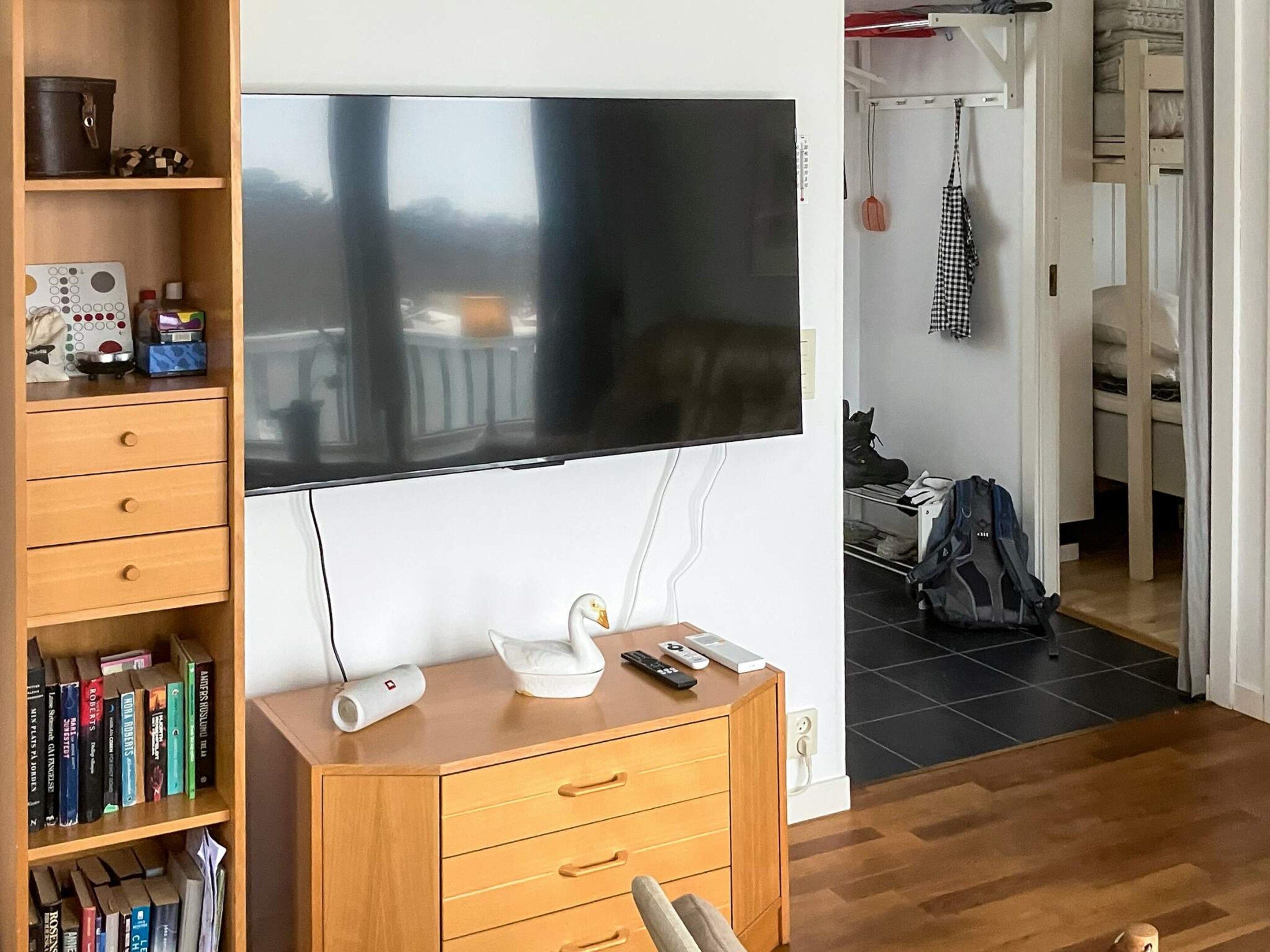 A room with a flat-screen TV mounted on the wall, a wooden cabinet with drawers, a bookshelf with books, and a doorway leading to another room.
