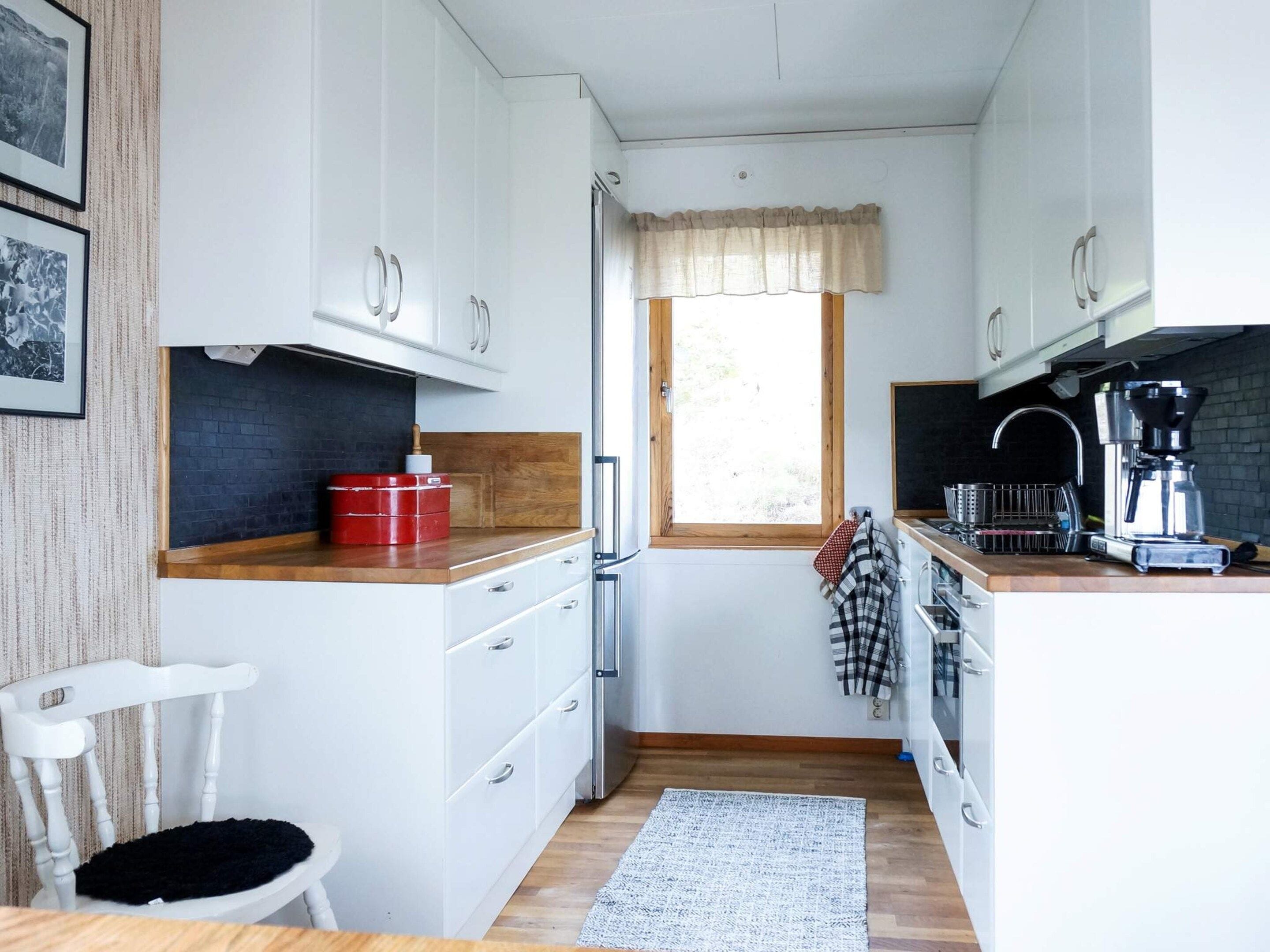A compact kitchen with white cabinets, a wooden countertop, a window with a valance, a coffee machine, and a black rug.