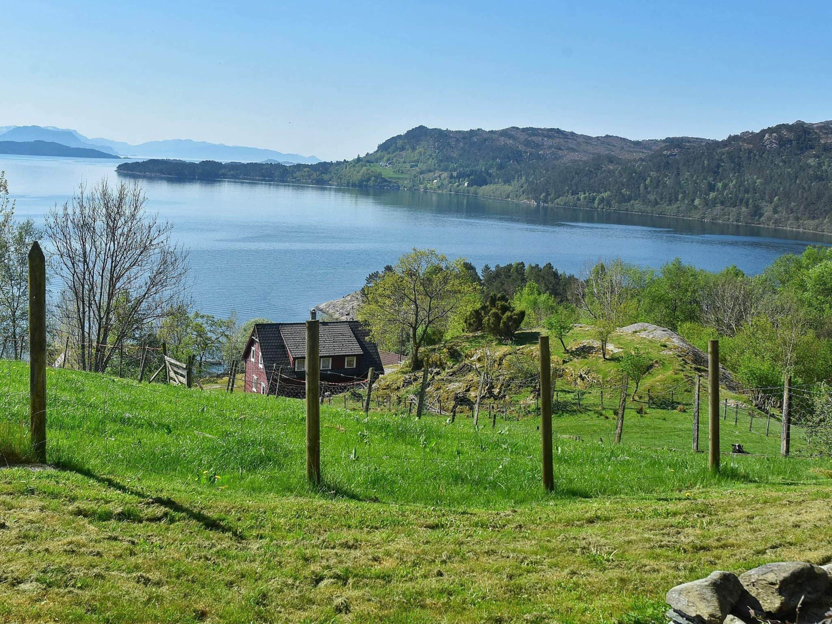 A house with a sloped roof, surrounded by a fenced yard with grass and trees, overlooking a large body of water and distant mountains.