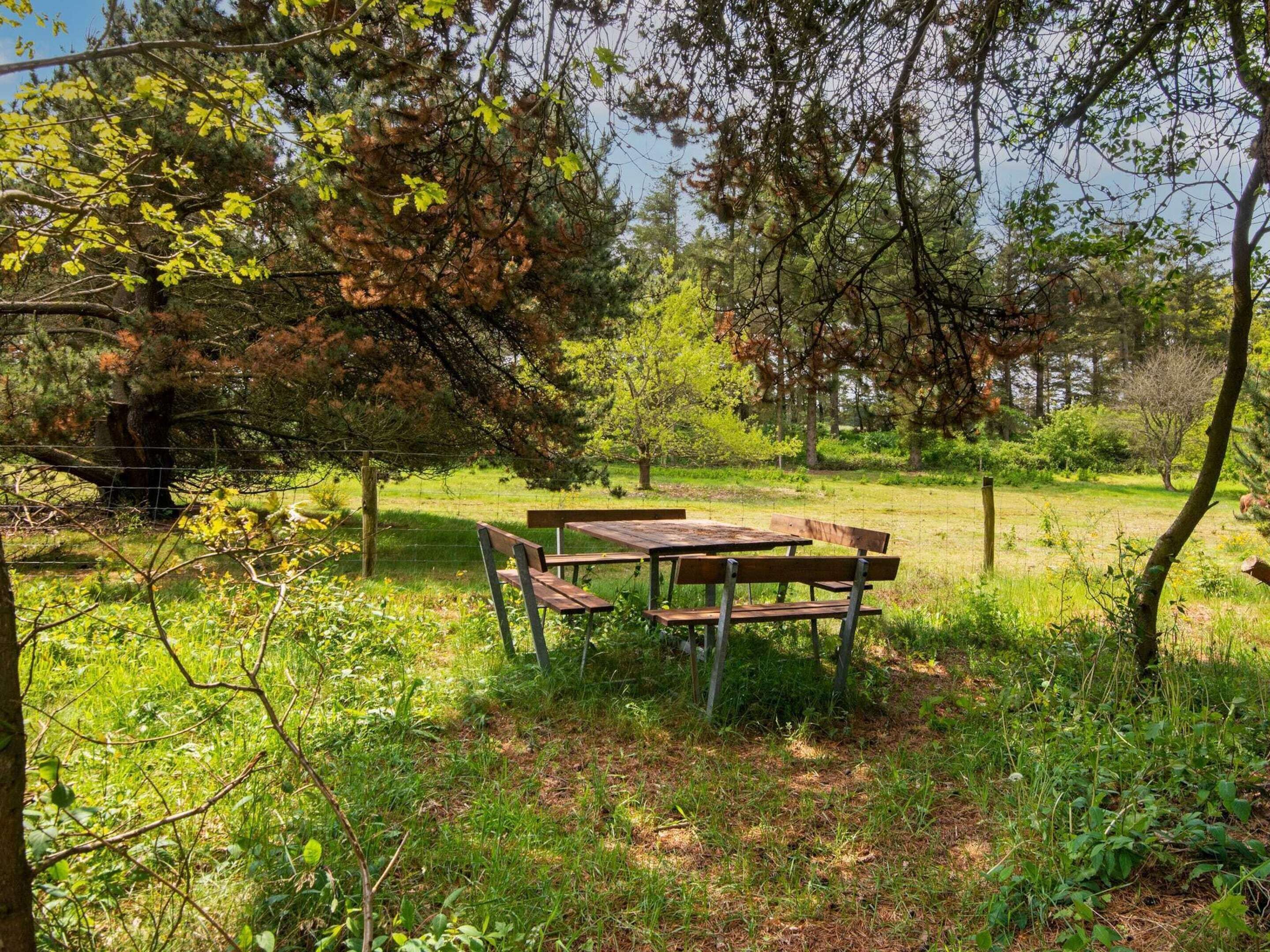 A wooden picnic table and benches in a grassy area with trees and a fence in the background.
