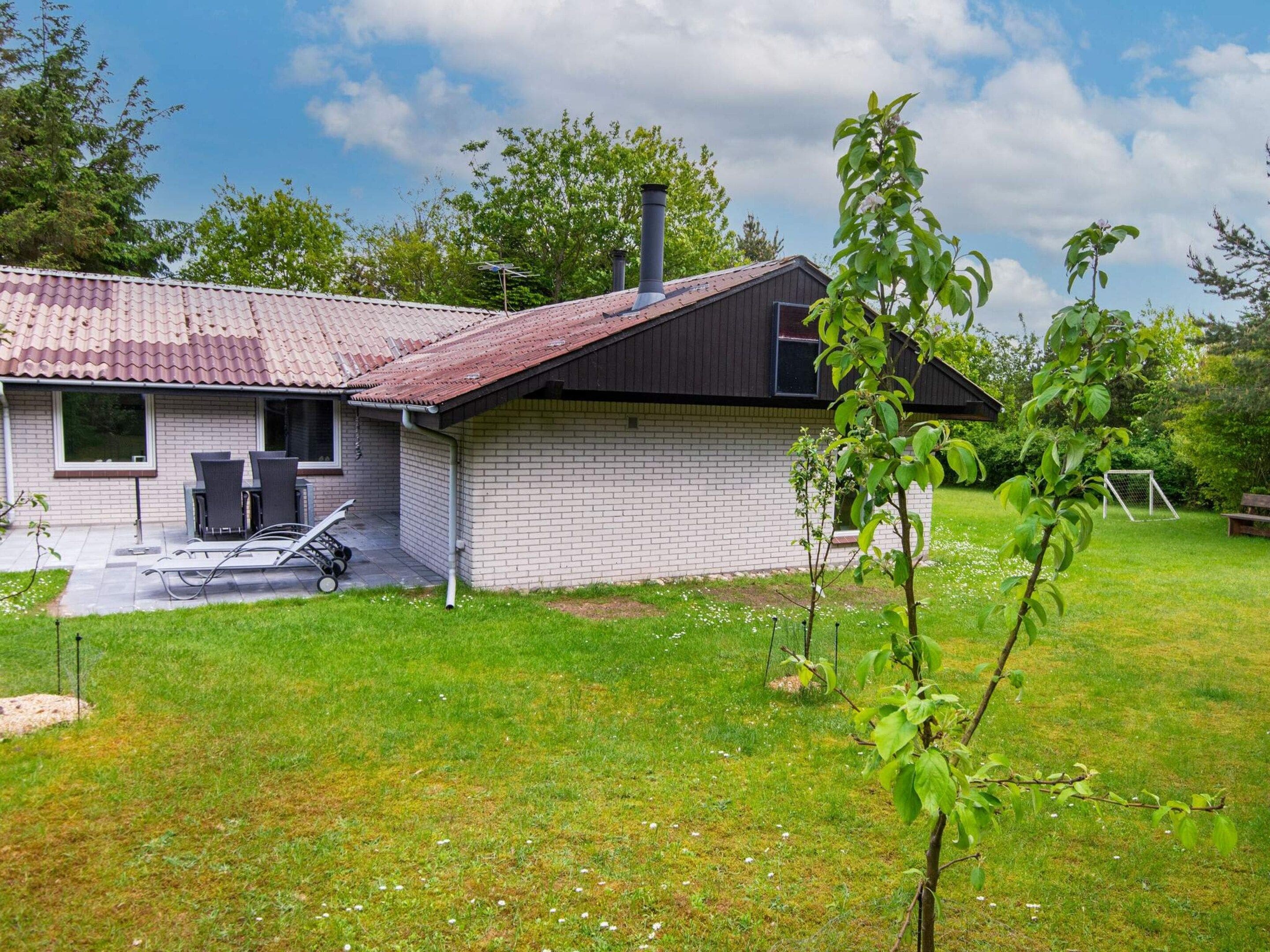 A single-story house with a tiled roof, a brick exterior, and a small front yard with a lawn and a young tree.