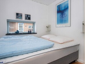A bedroom with a bed, a window with blinds, a wall-mounted light, and two framed artworks.