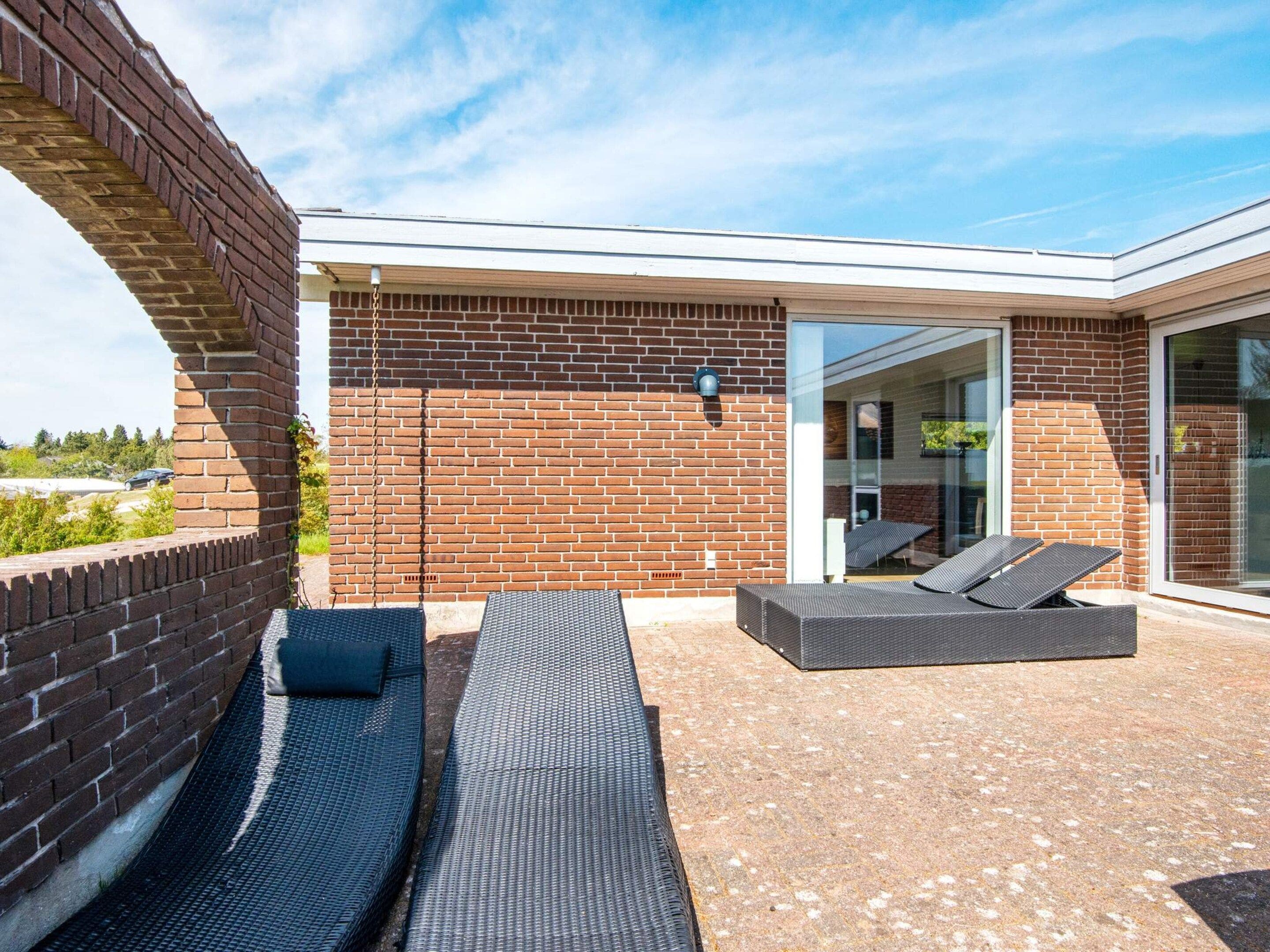 Une terrasse extérieure moderne, avec des murs en briques, une porte d’entrée en verre et deux fauteuils de couleur sombre.