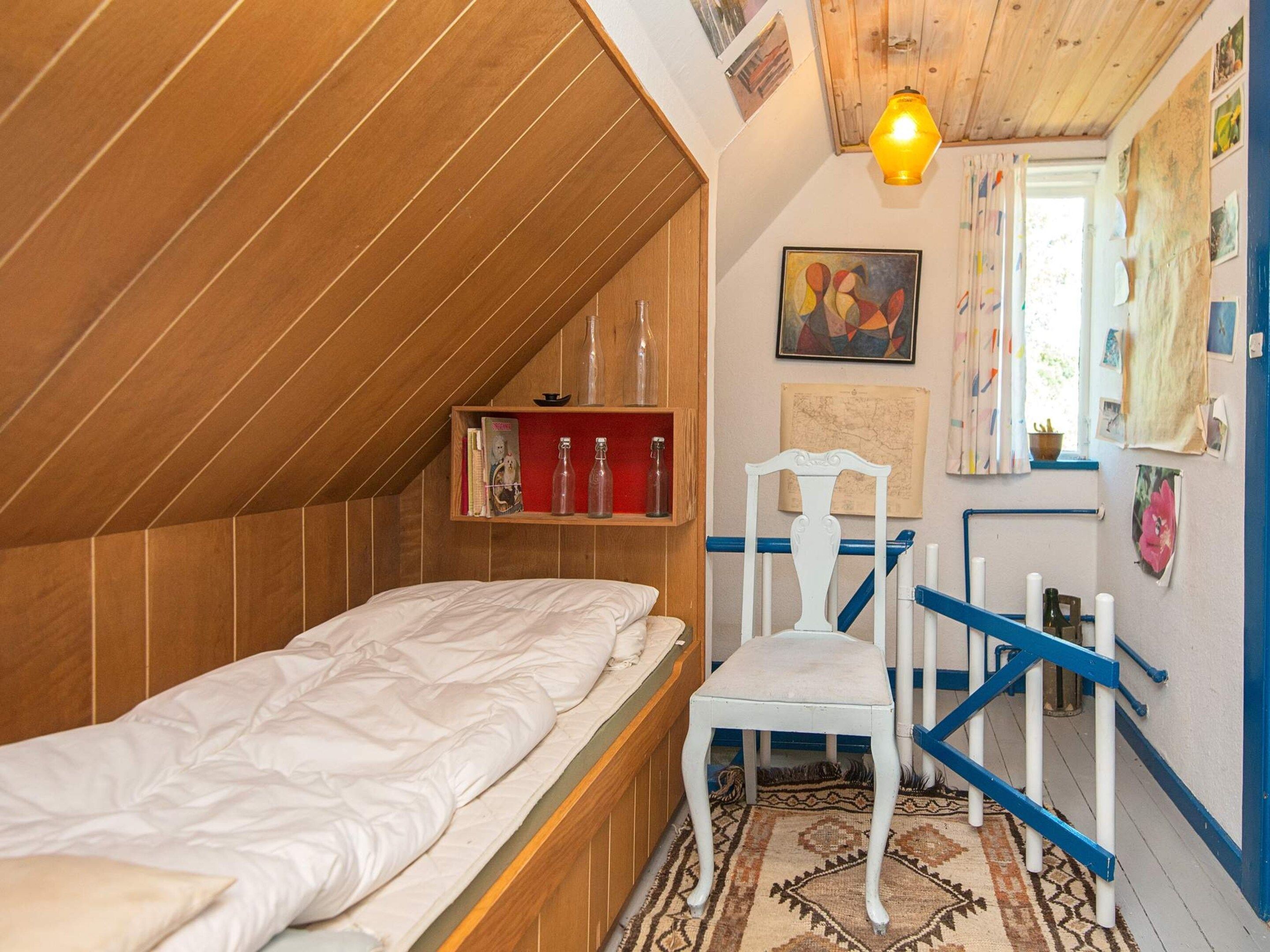 A bedroom with a sloped ceiling, a bed, a chair, and a shelf with books and bottles.