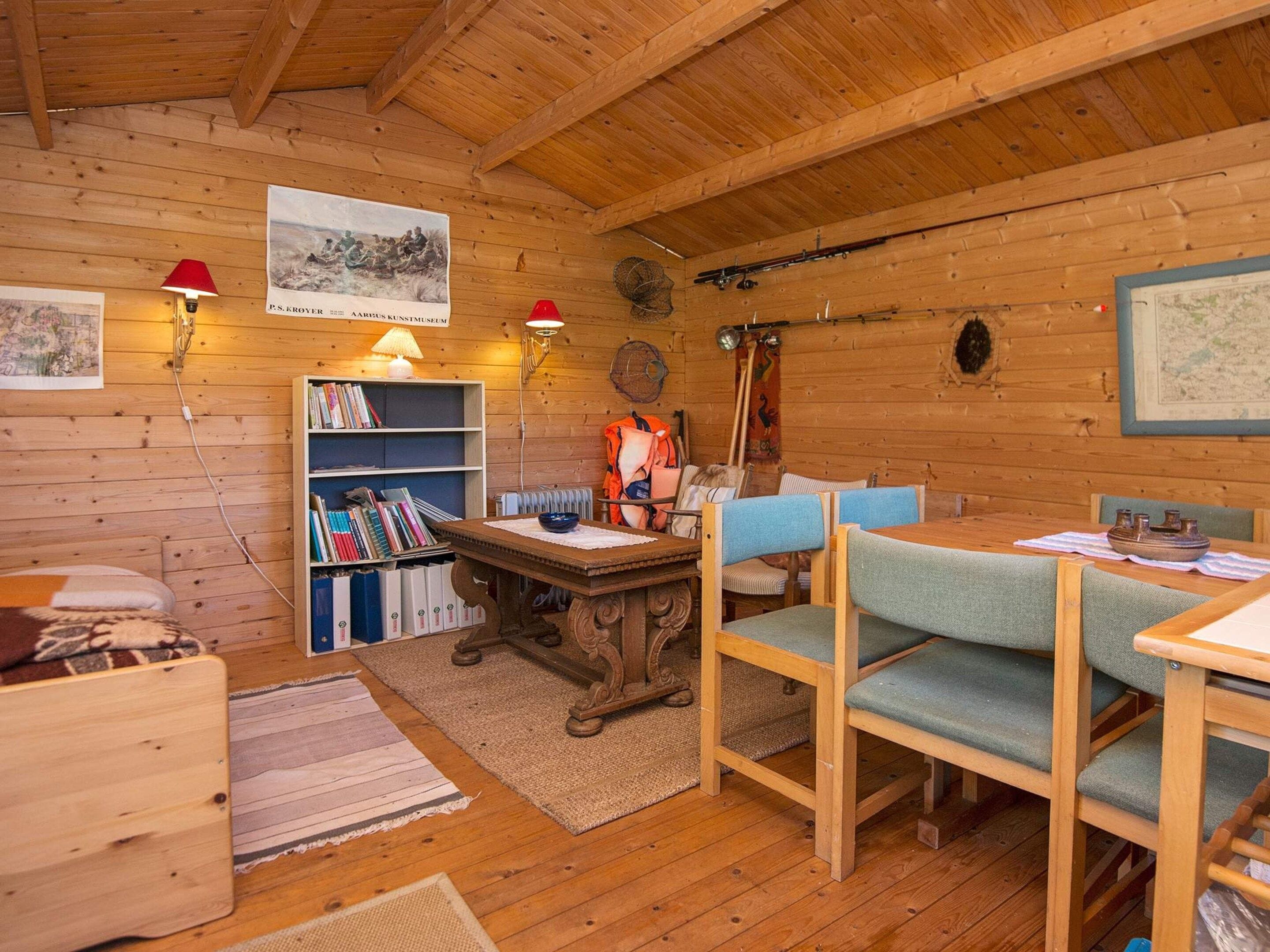 A wooden cabin interior with a dining table, chairs, a bookshelf, and wall-mounted lamps.