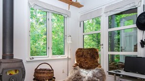 Una habitación acogedora con una estufa de leña, ventanas amplias con vistas a la vegetación, un sillón cómodo y un televisor sobre un mueble.