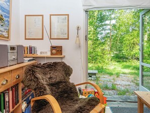 A cozy room with a fur chair, wooden shelves with books, a radio, and a view of a green garden through a glass door. - 4 Person Holiday Home in Bryrup (Bryrup)