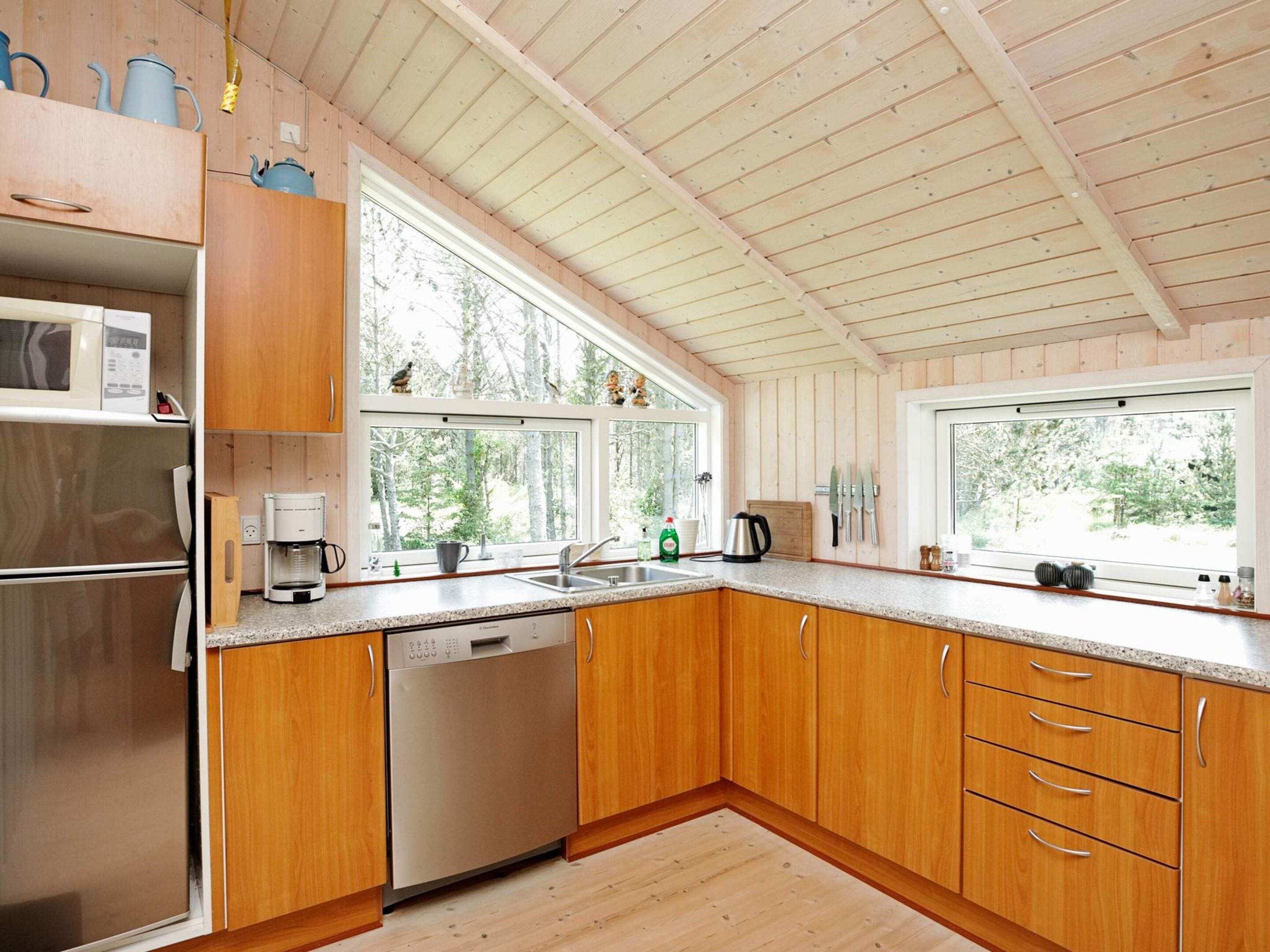 Une cuisine moderne avec des meubles en bois, un réfrigérateur en acier inoxydable, un lave-vaisselle et de grandes fenêtres offrant une vue sur la forêt.