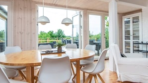 Een moderne eetruimte met een houten tafel en witte stoelen, grote ramen en uitzicht op groen.