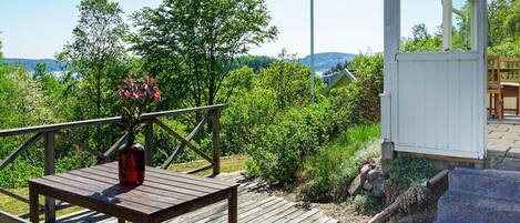 A wooden deck with a table and chairs, a glass enclosure, and a view of greenery and hills.