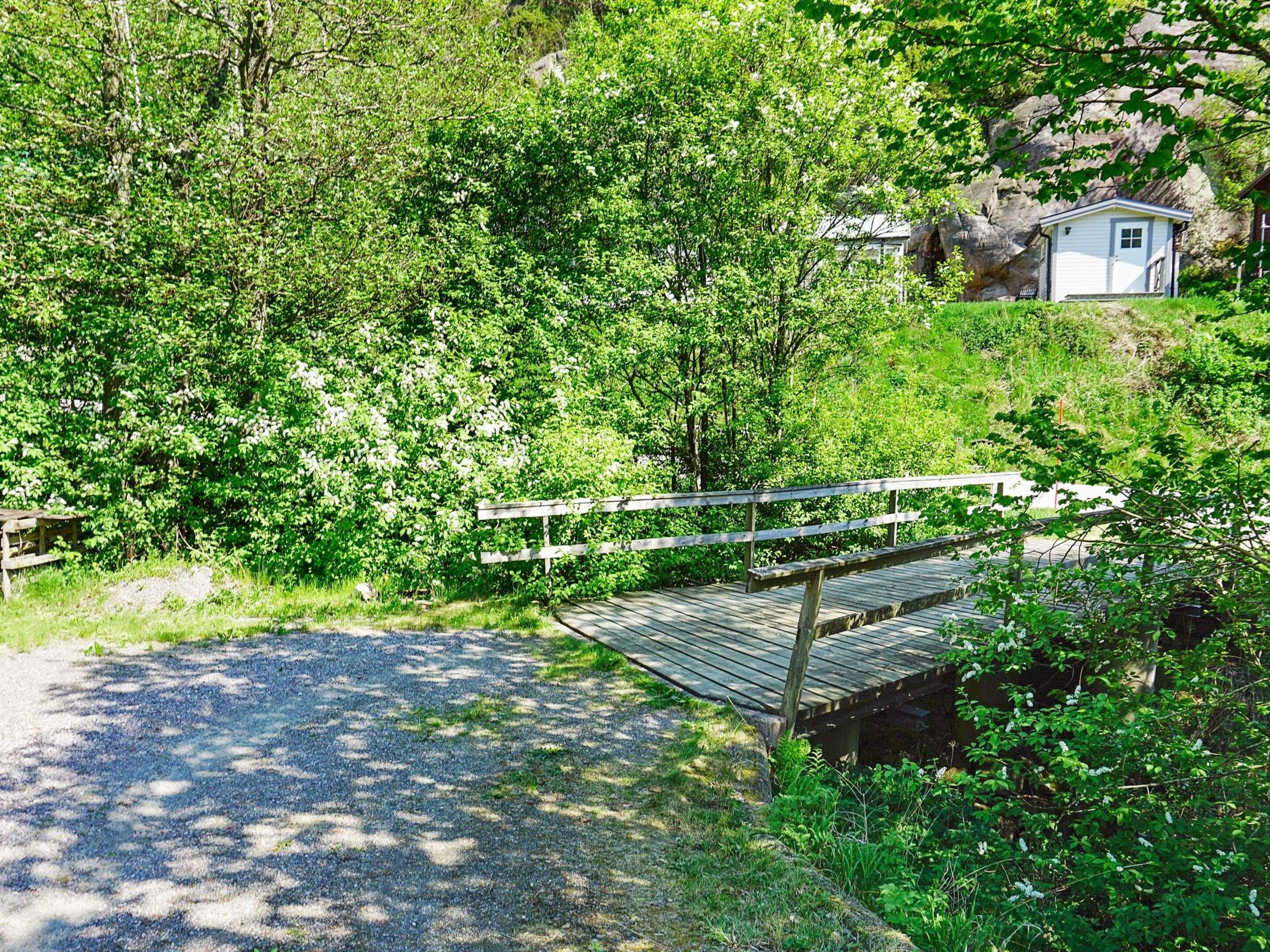 En grussti fører hen til en træterrasse med gelænder, omgivet af frodig vegetation og en bygning i baggrunden.