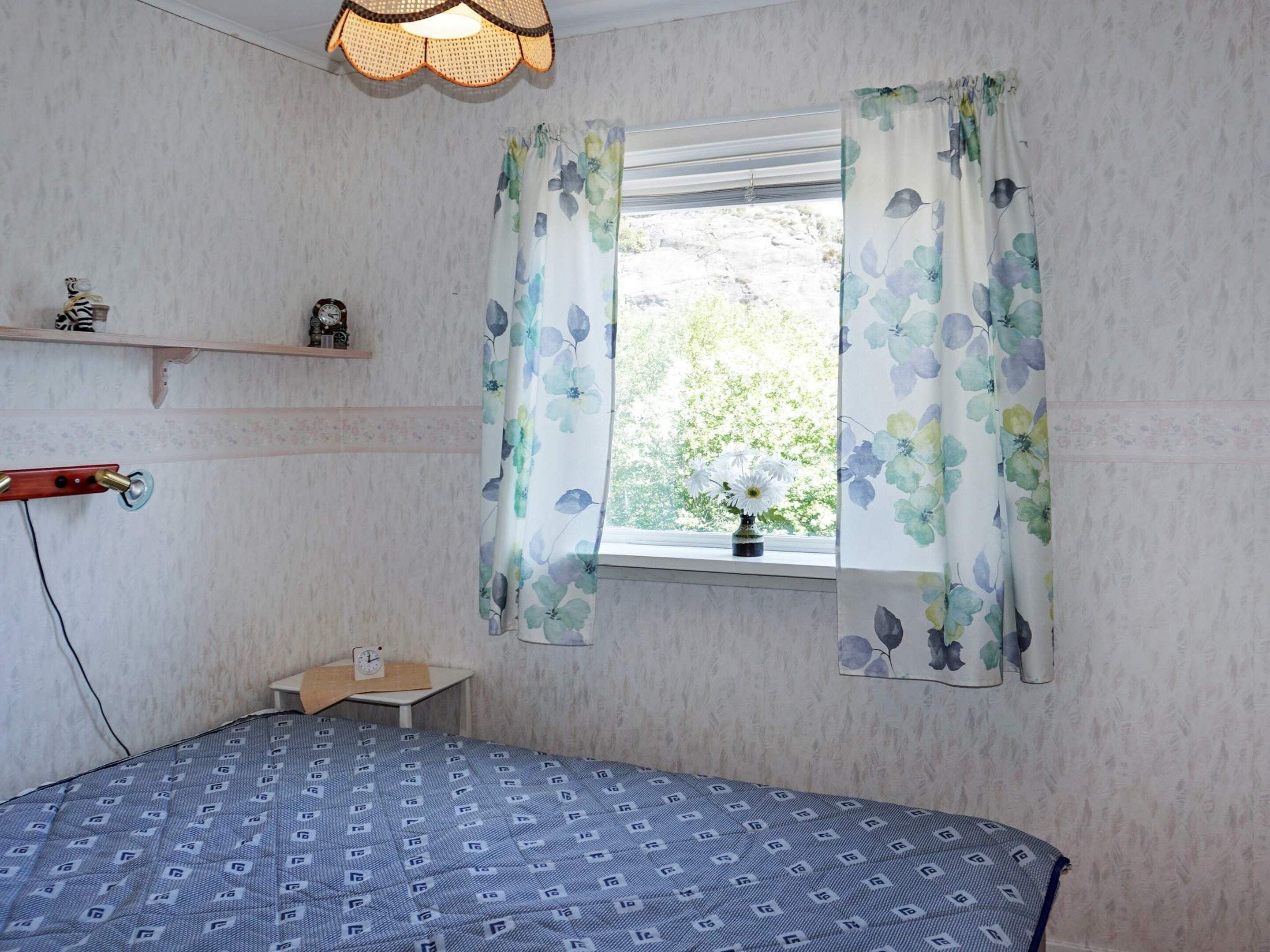 Quarto com cama, janela com cortinas florais, mesa de cabeceira com relógio e prateleira com objetos decorativos.