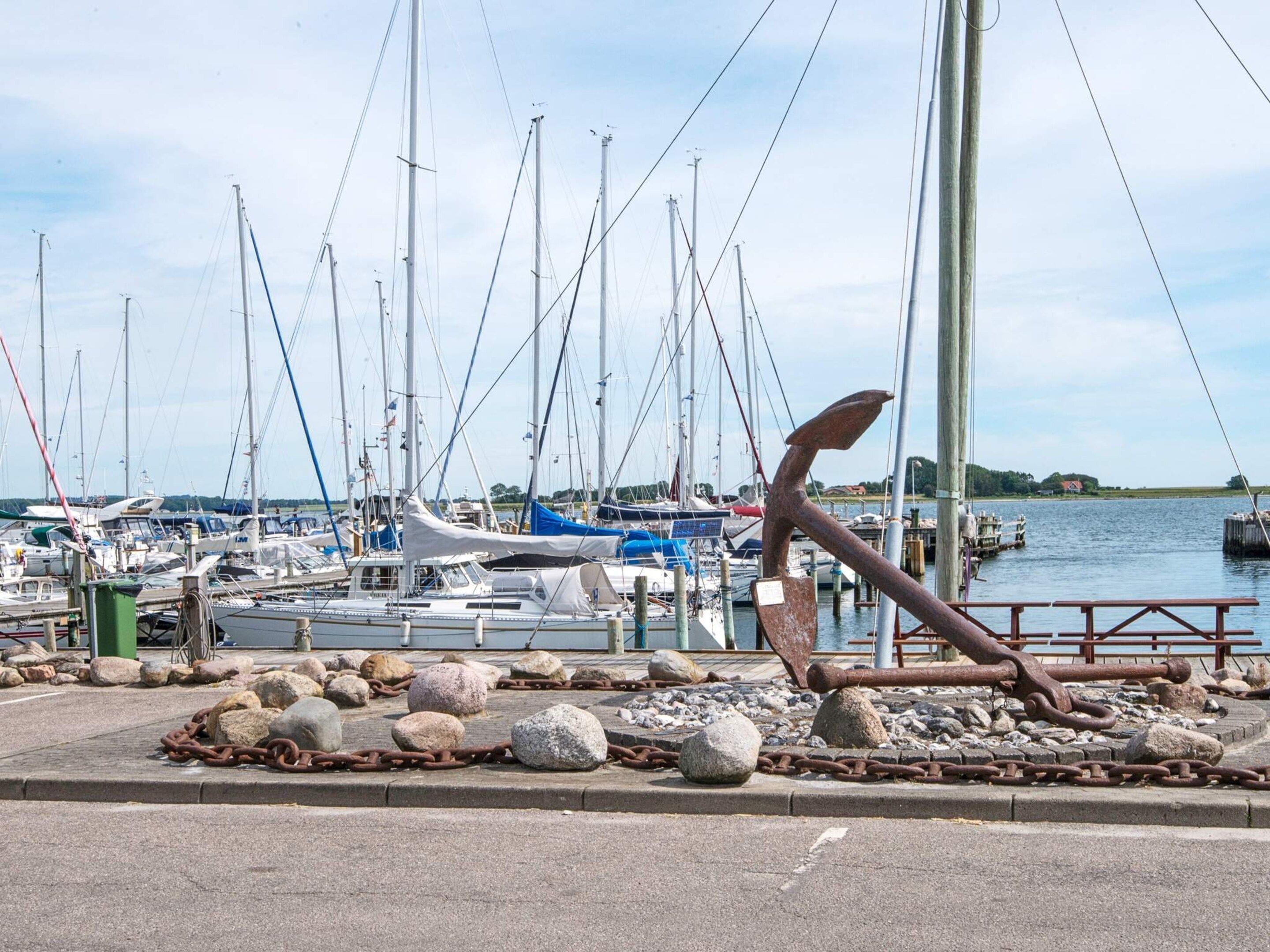 Un puerto deportivo con barcos amarrados, una gran escultura de ancla y una masa de agua tranquila.