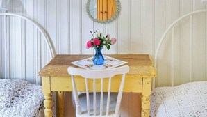 A room with a wooden table, a white chair, and two beds with lace covers.