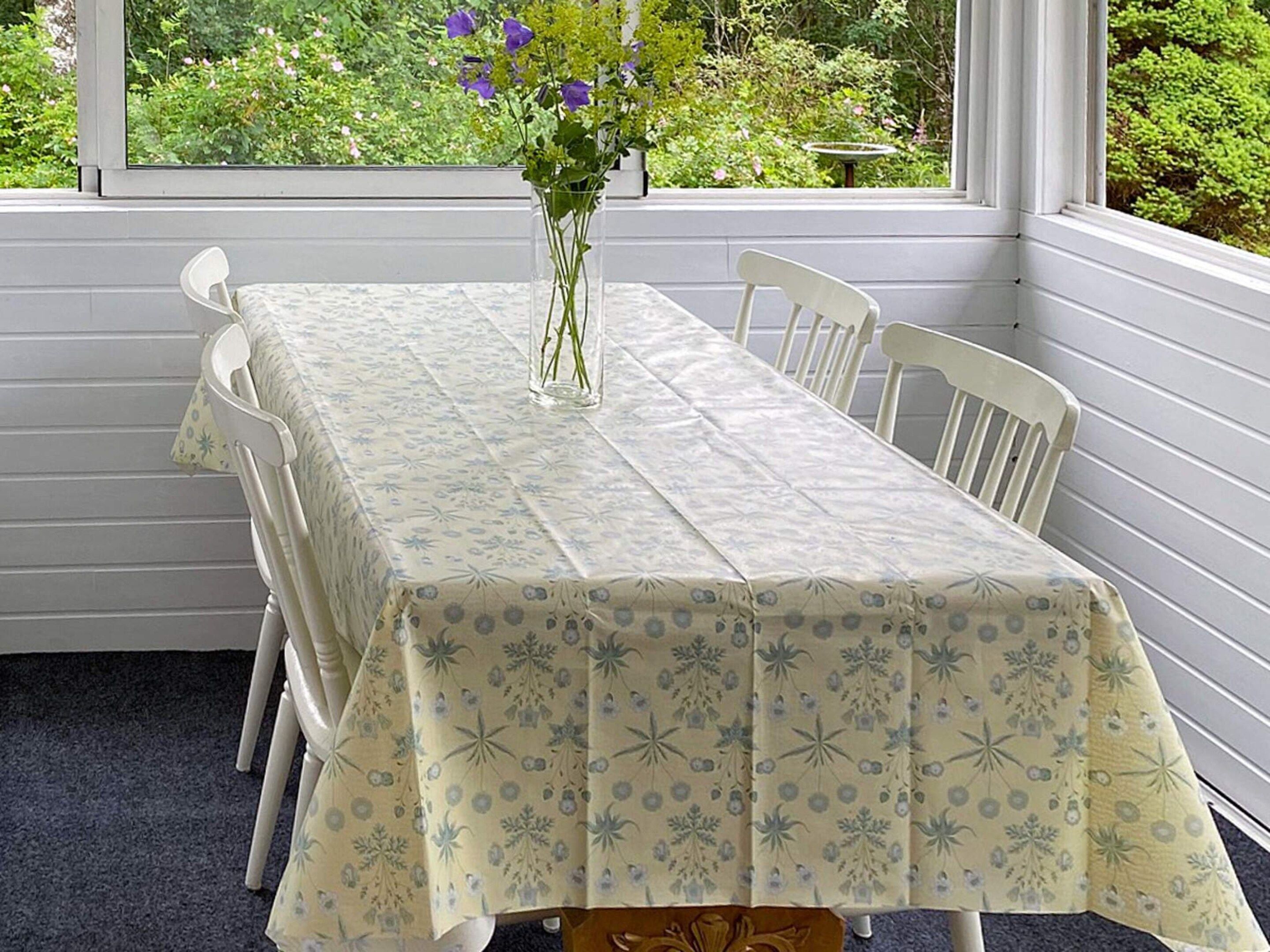 A dining table with a floral tablecloth, surrounded by white chairs, set against a window with a view of greenery.