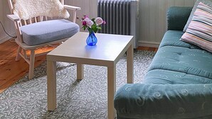 A room with a light-colored armchair, a blue vase with flowers, a small white table, a radiator, and a teal sofa with patterned cushions.
