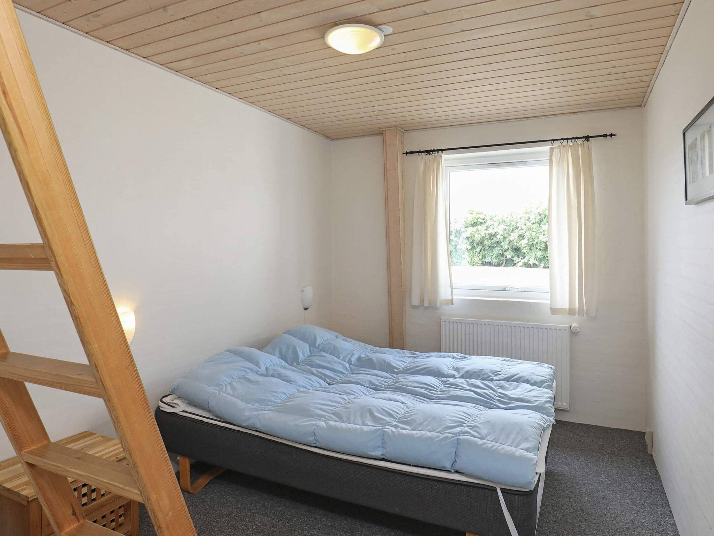 A single bed with a blue mattress in a room with a wooden ceiling, a window with curtains, and a radiator.