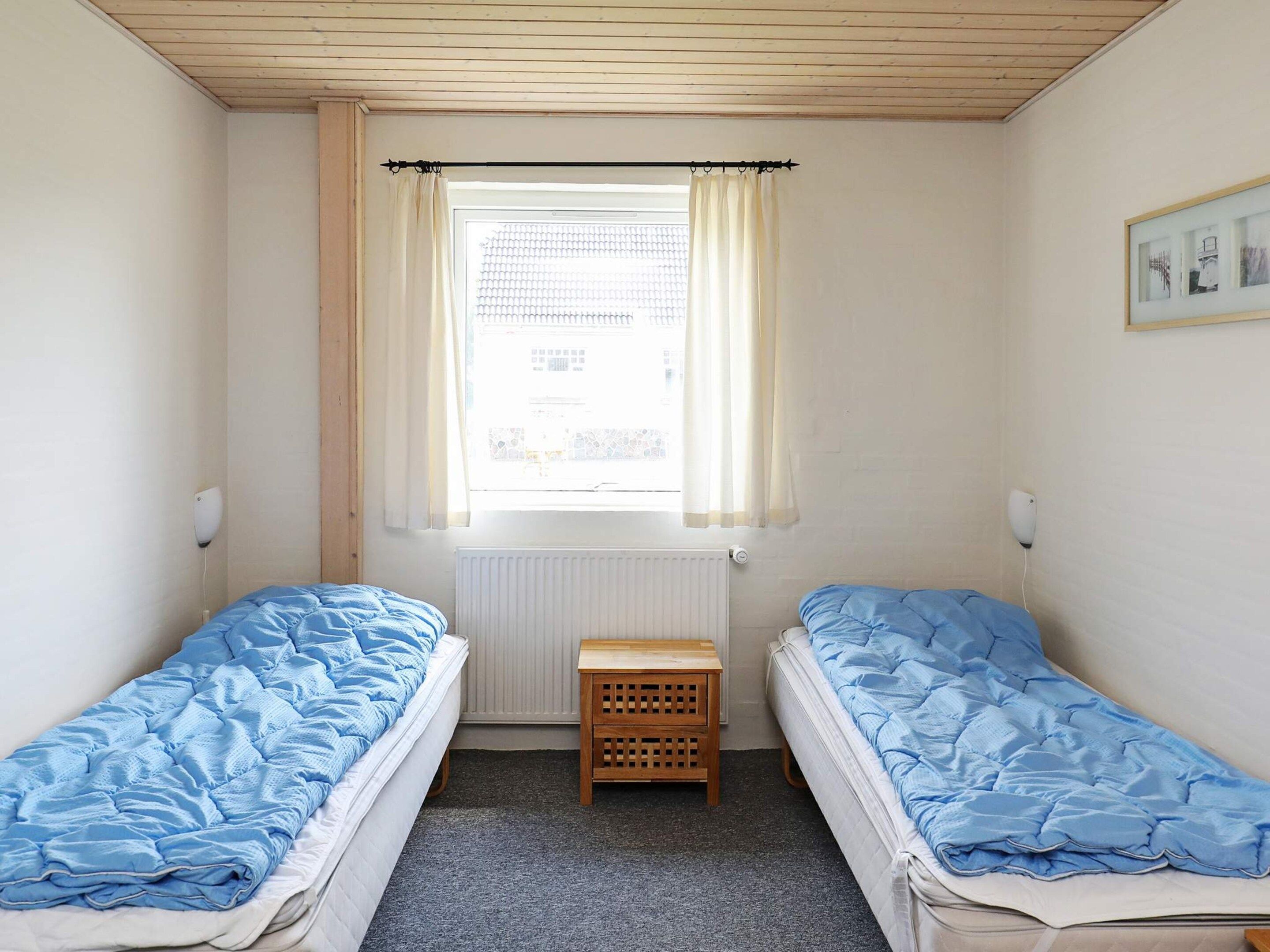 A small room with two single beds, a wooden nightstand, a radiator, and a window with curtains.