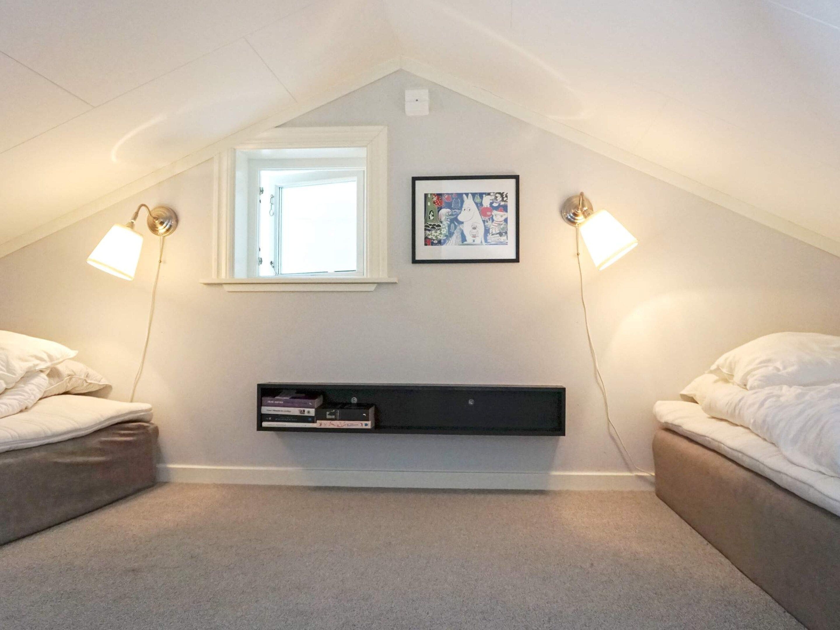 Une petite chambre mansardée avec deux lits, une fenêtre, une étagère remplie de livres et des lampes fixées au mur.