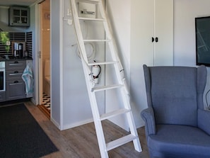 A white ladder leaning against a wall, a grey armchair, and a kitchen area with a stove and cabinets.