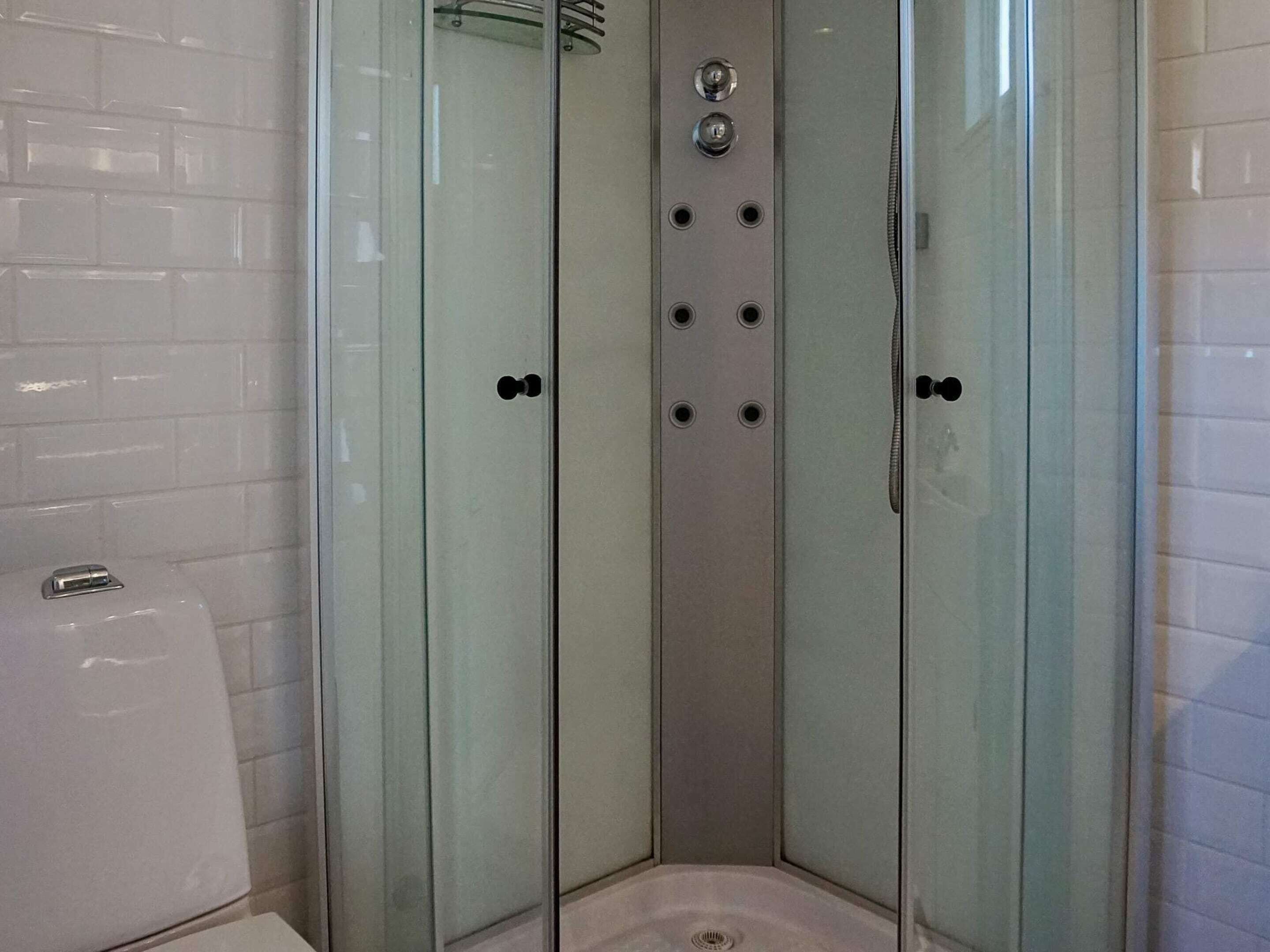 Une cabine de douche fermée par des parois en verre, avec un mur recouvert de carreaux blancs, un drain visible et une douchette à main.