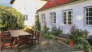 A white building with a red-tiled roof, a wooden table with chairs, and a garden with flowers and plants.