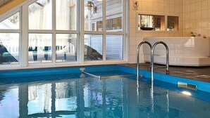 An indoor swimming pool with clear glass walls, a blue pool edge, and a white tiled wall with a bathtub. - Holiday Home in Auklandshamn (Sveio)
