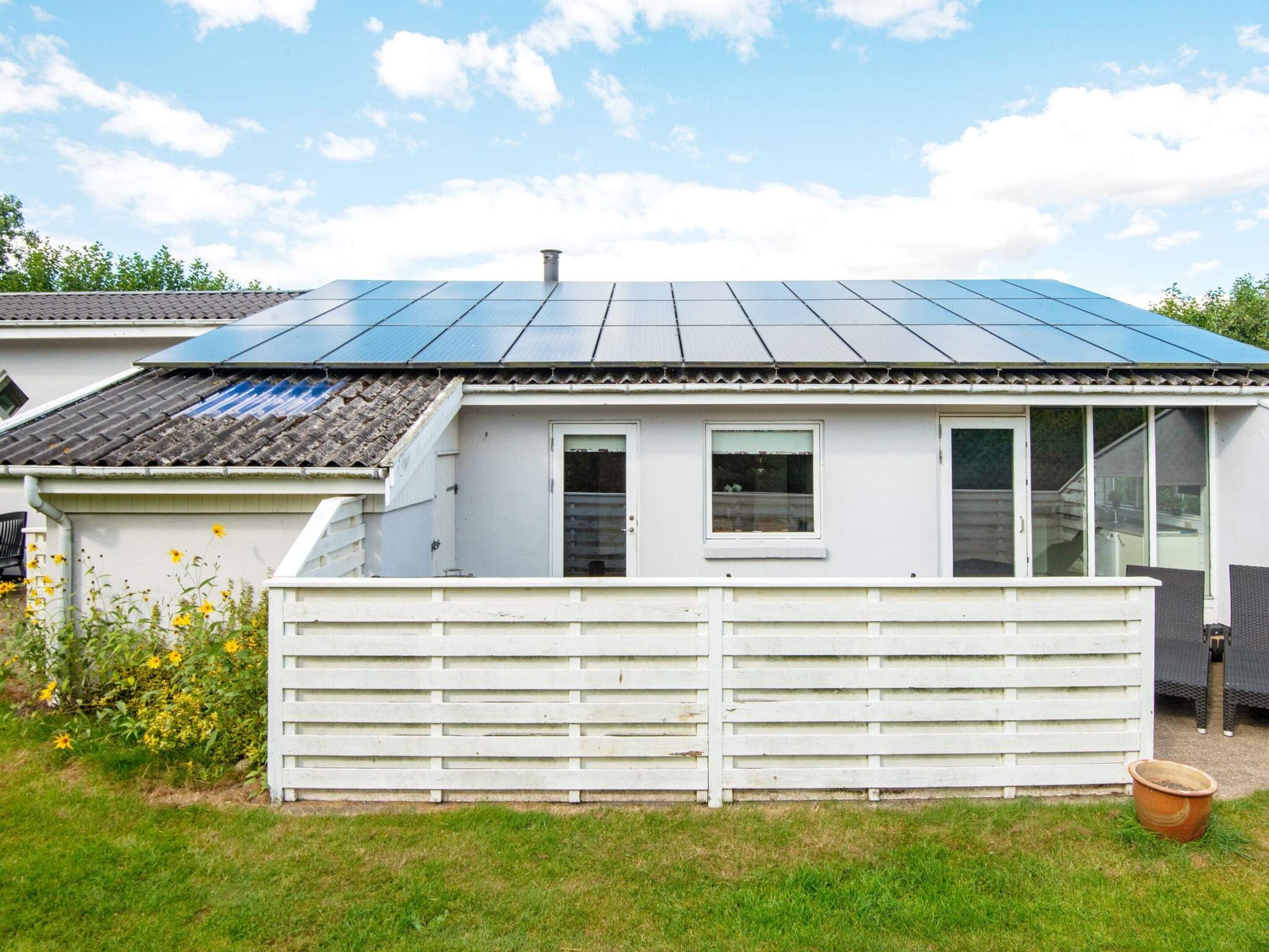 A house with a solar panel roof, a white fence, and a garden with yellow flowers.
