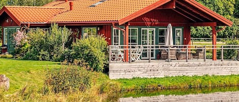 Una cabaña de madera con techo rojo, un porche cubierto con sillas y un reflejo en el agua.