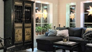Een gezellige woonkamer met een donkerblauwe bank, een bijpassend poufje en een houten salontafel. Een schommelstoel staat bij een grote kast met ingewikkelde decoraties. De kamer heeft grote ramen, waardoor ster-achtige versieringen zichtbaar zijn.
