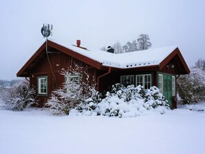 A wooden cabin with a sloped roof and a chimney, surrounded by snow-covered vegetation.