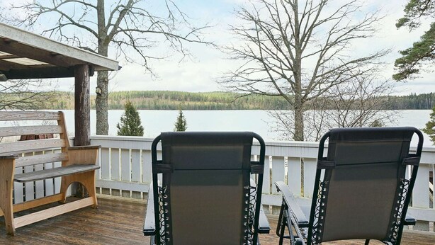 A wooden deck with two black chairs and a bench, overlooking a lake with trees and a cloudy sky. - 4 Person Holiday Home in Vetlanda (Vetlanda)