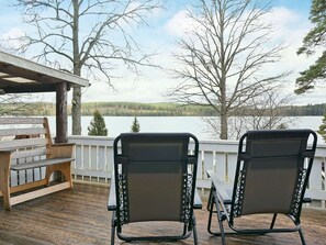 A wooden deck with two black chairs and a bench, overlooking a lake with trees and a cloudy sky. - 4 Person Holiday Home in Vetlanda (Vetlanda)