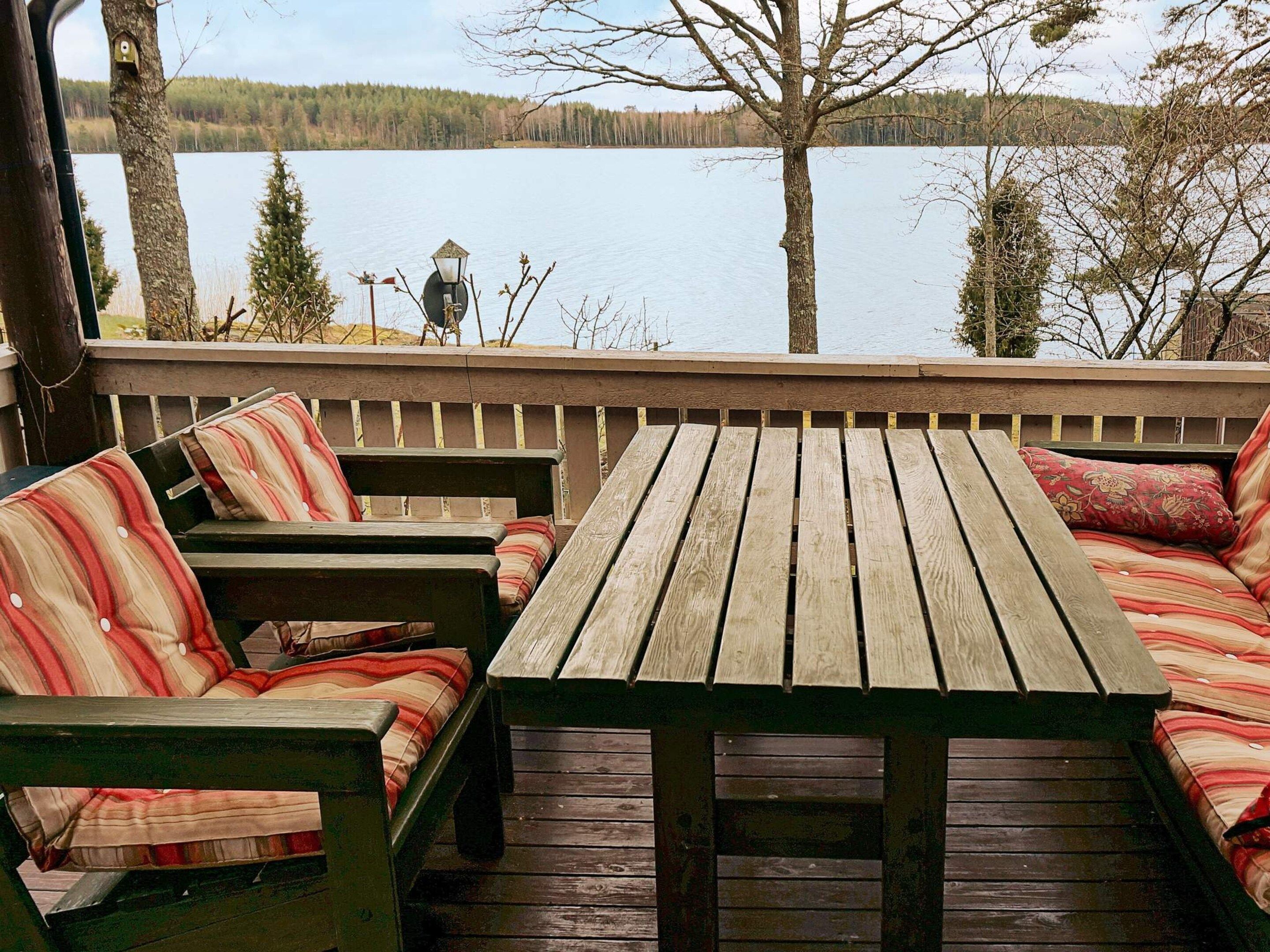 Een houten terras met een tafel en zitplaatsen met kussens, met uitzicht op een kalm meer met bomen op de achtergrond.