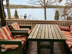 A wooden deck with a table and cushioned seating, overlooking a calm lake with trees in the background. - 4 Person Holiday Home in Vetlanda (Vetlanda)
