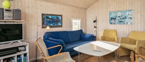 A cozy living room with a blue sofa, yellow armchairs, a white coffee table, and a television on a wooden shelf.