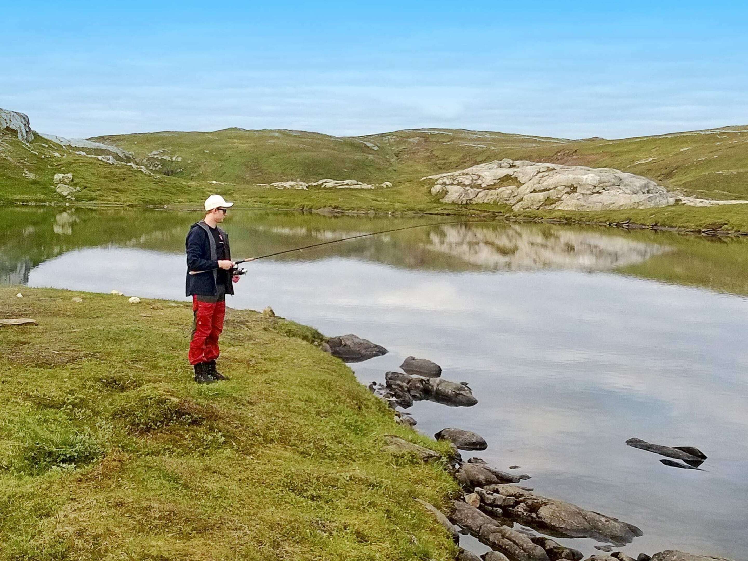 Una persona pescando en un lago tranquilo con orillas cubiertas de hierba y afloramientos rocosos.